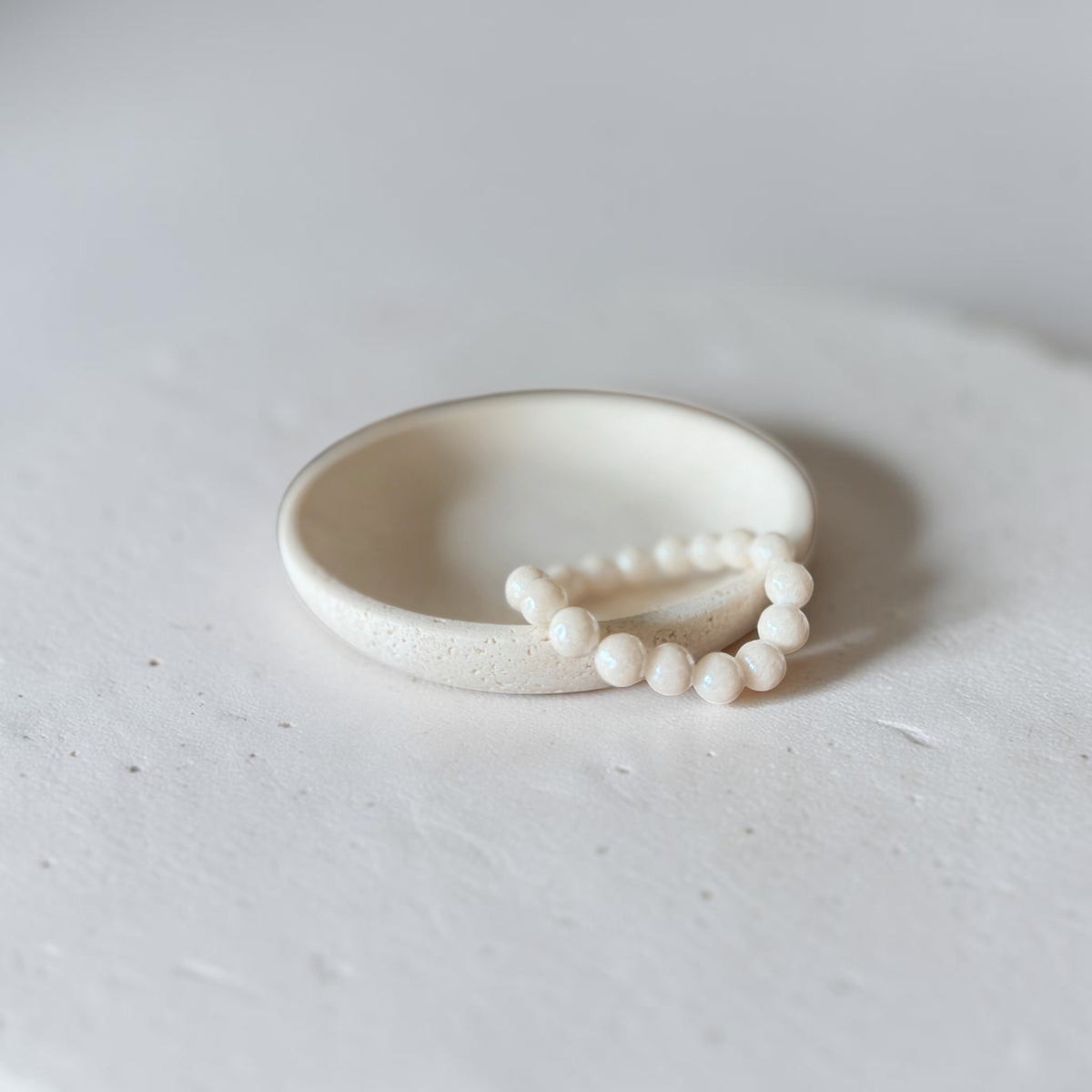 A minimalist cream-colored ceramic ring with a small beaded accent, photographed on a textured light gray surface.