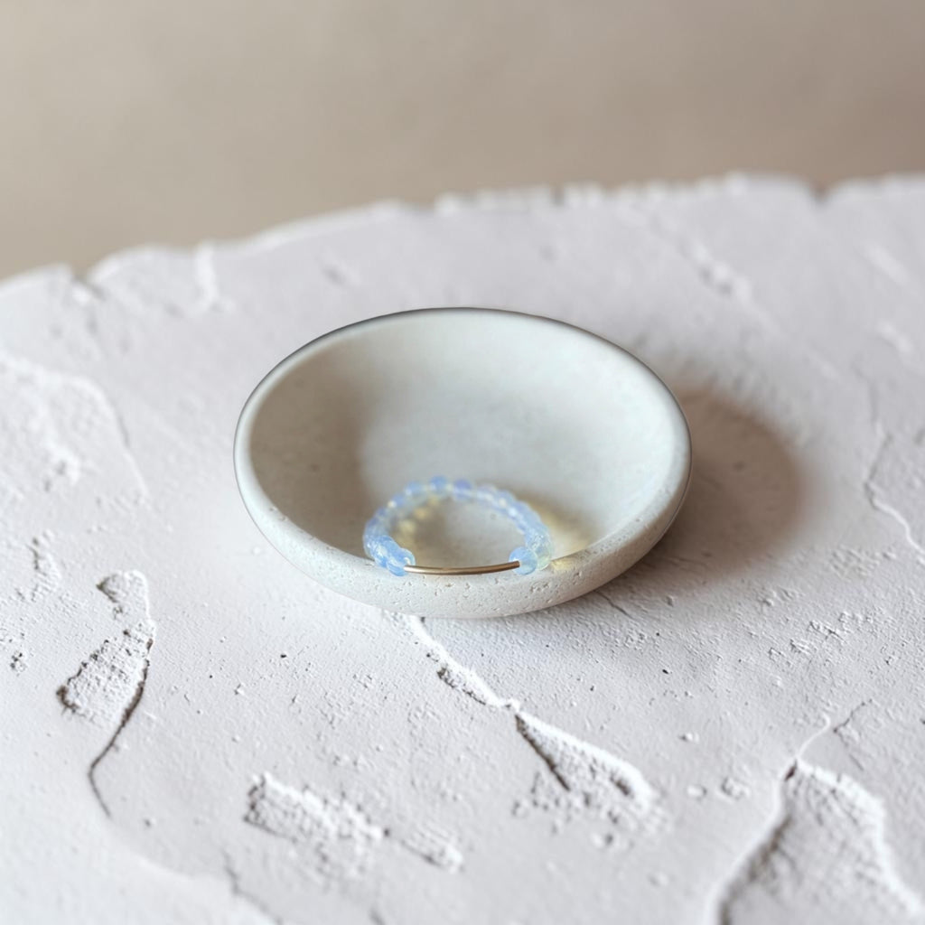 A small handmade ceramic dish with a light beige glaze and a thin dark rim, holding a delicate light blue beaded ring, set on a textured white surface.