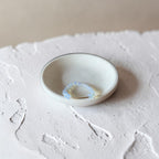 A small handmade ceramic dish with a light beige glaze and a thin dark rim, holding a delicate light blue beaded ring, set on a textured white surface.
