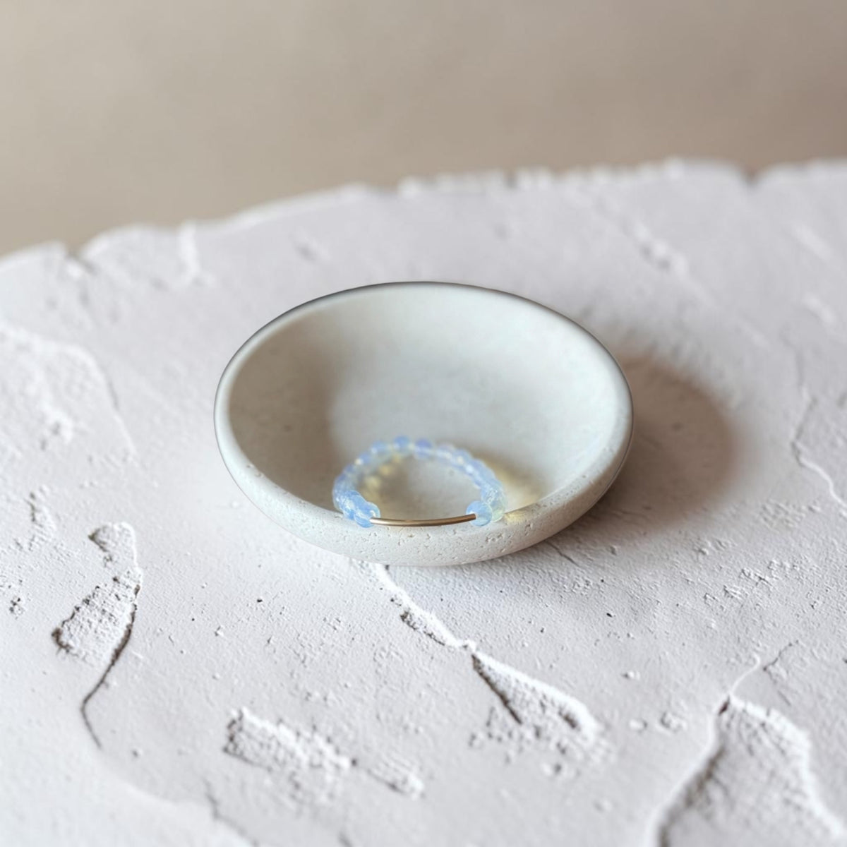 A small handmade ceramic dish with a light beige glaze and a thin dark rim, holding a delicate light blue beaded ring, set on a textured white surface.