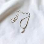 A delicate necklace with a small iridescent shell pendant, featuring a gold-toned chain with a black spacer bead and a textured silver-toned section, laid on a soft white fabric background.