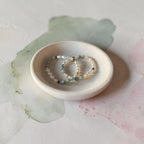 Two beaded stretch bracelets with light blue, white, and dark beads resting in a small round ceramic dish on a textured light background with soft watercolor washes.