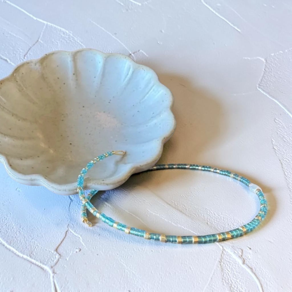 A beaded bracelet with turquoise blue, yellow, and white beads, displayed on a textured white surface next to a small ceramic dish.