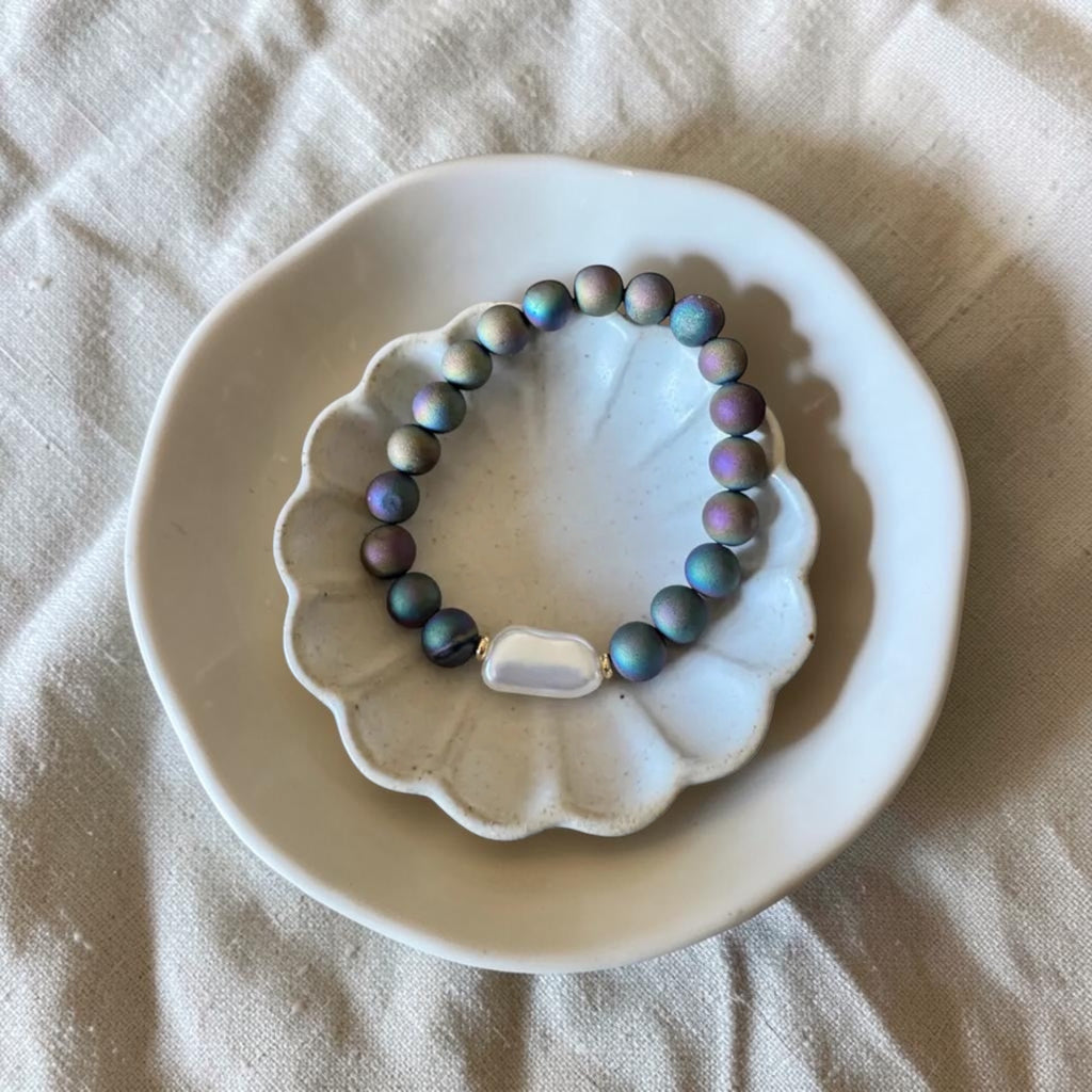 A beaded bracelet with iridescent blue, purple, and green beads, featuring a white shell-shaped bead in the center, displayed on a white scalloped dish over a beige fabric background.