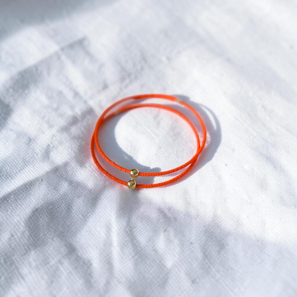 A pair of thin orange wire bracelets with small gold bead accents, resting on a textured white fabric background.