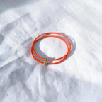 A pair of thin orange wire bracelets with small gold bead accents, resting on a textured white fabric background.