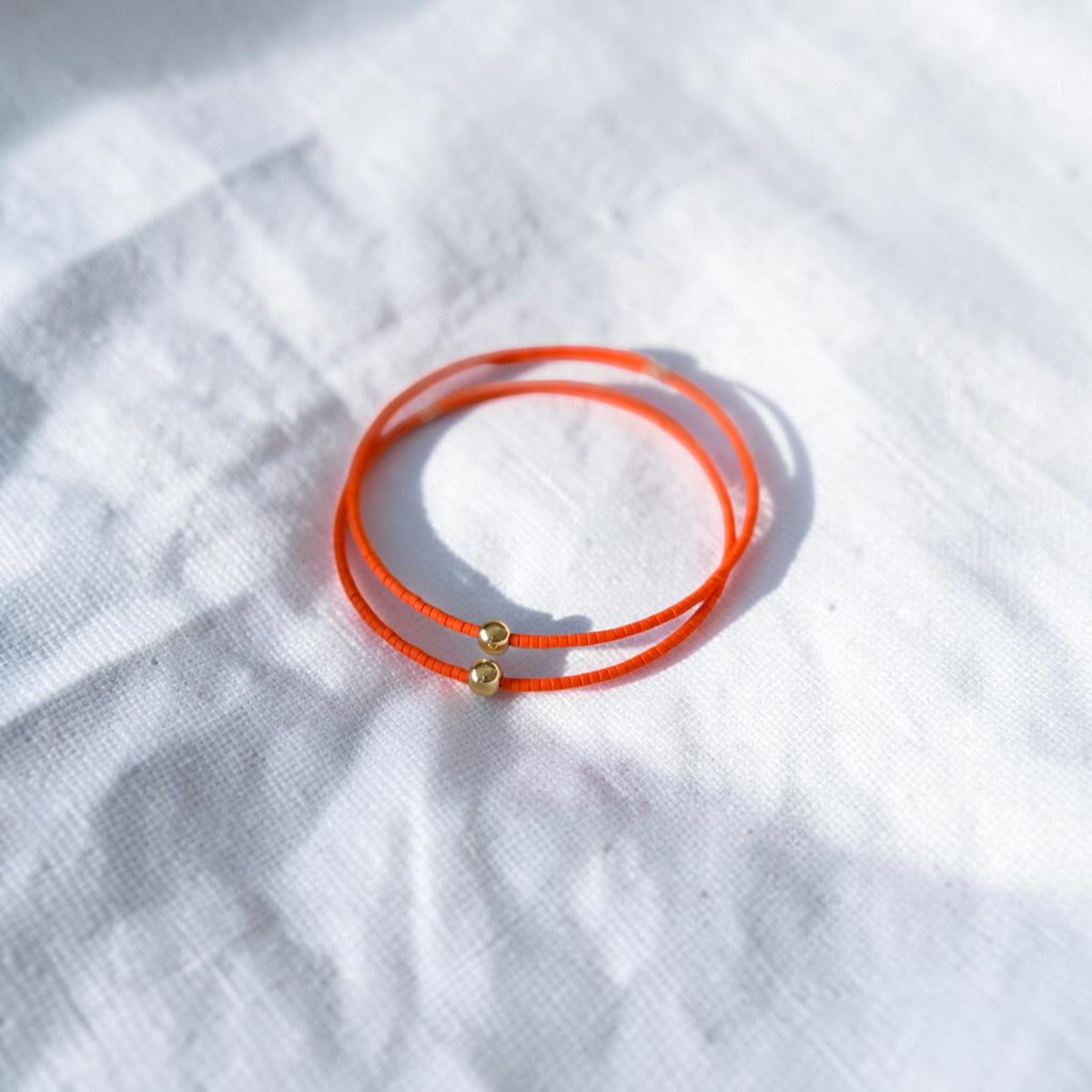 A pair of thin orange wire bracelets with small gold bead accents, resting on a textured white fabric background.