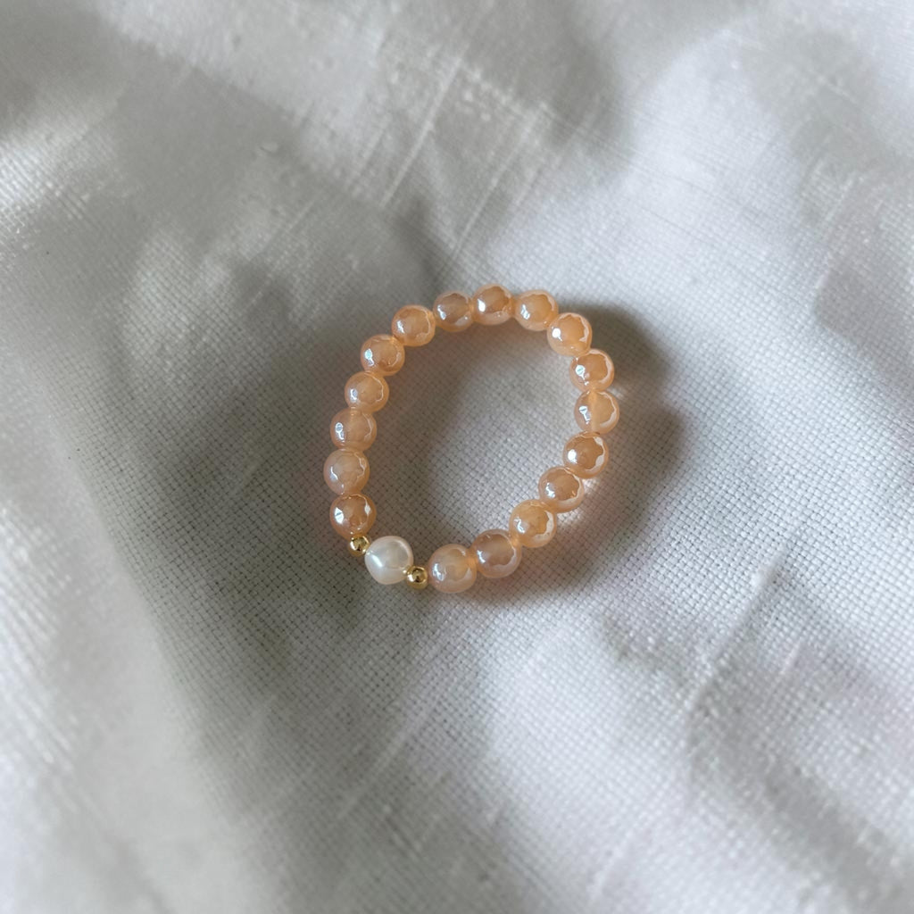 A beaded stretch bracelet featuring peach-colored round beads, a single white pearl, and a small gold spacer, resting on a textured white fabric background.
