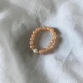 A beaded stretch bracelet featuring peach-colored round beads, a single white pearl, and a small gold spacer, resting on a textured white fabric background.
