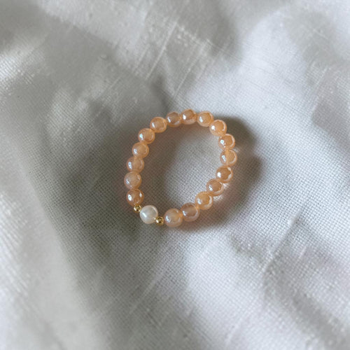 A beaded stretch bracelet featuring peach-colored round beads, a single white pearl, and a small gold spacer, resting on a textured white fabric background.