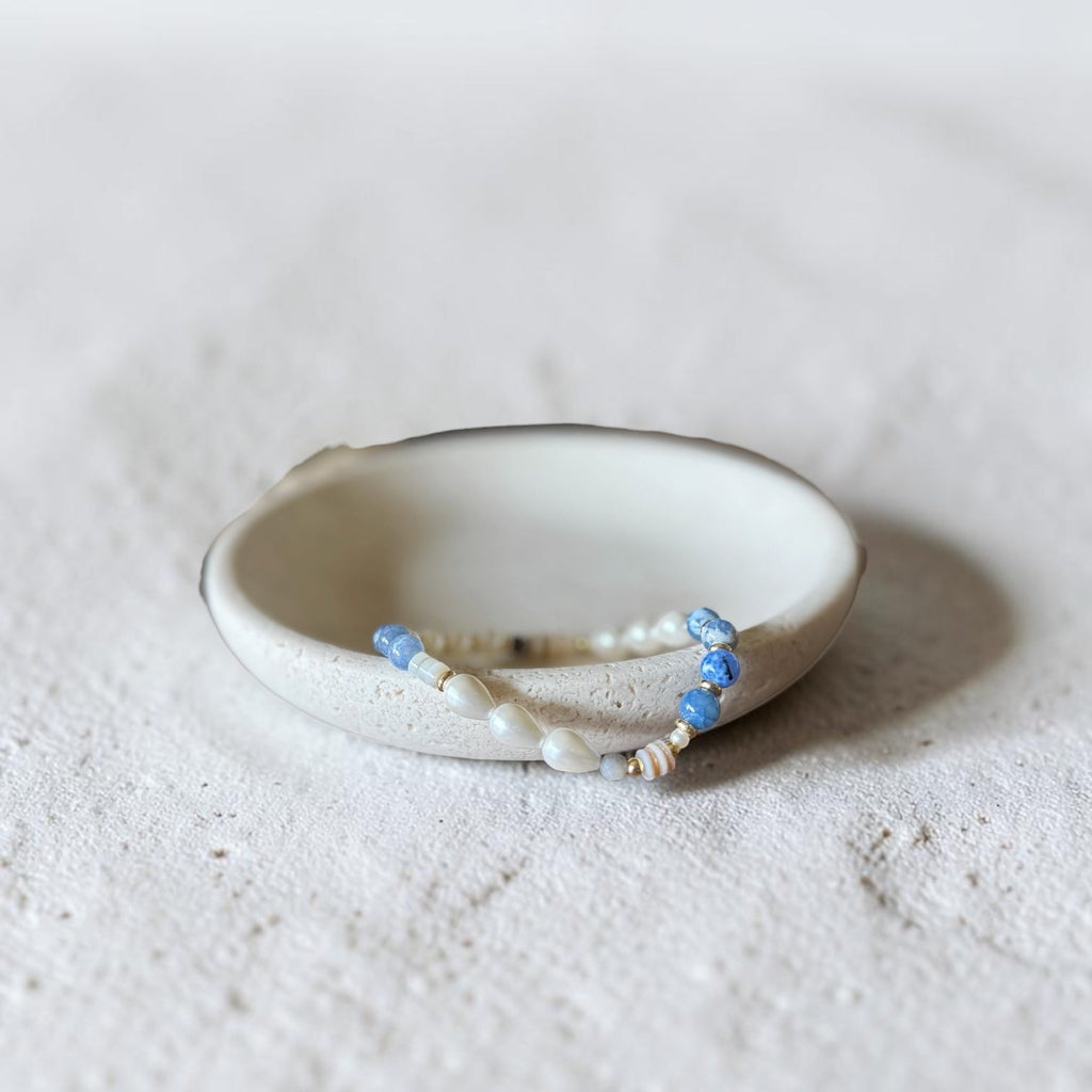 A handmade ceramic holder with a beaded bracelet resting inside it, set against a textured light gray background.