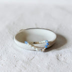 A handmade ceramic holder with a beaded bracelet resting inside it, set against a textured light gray background.