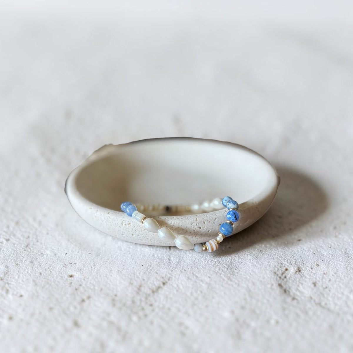 A handmade ceramic holder with a beaded bracelet resting inside it, set against a textured light gray background.