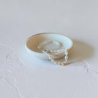 A delicate strand of white and cream pearl bracelets draped over a small, shallow, textured ceramic dish on a light gray surface.