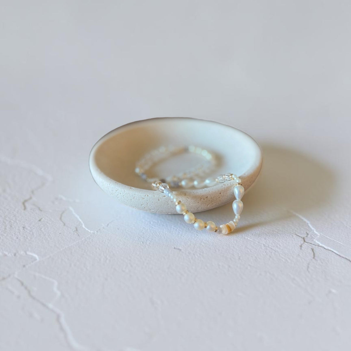 A delicate strand of white and cream pearl bracelets draped over a small, shallow, textured ceramic dish on a light gray surface.