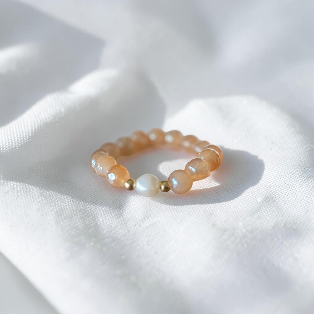 A beaded stretch bracelet featuring translucent peach-orange beads, a white pearl, and small gold spacer beads, resting on a textured white fabric background.