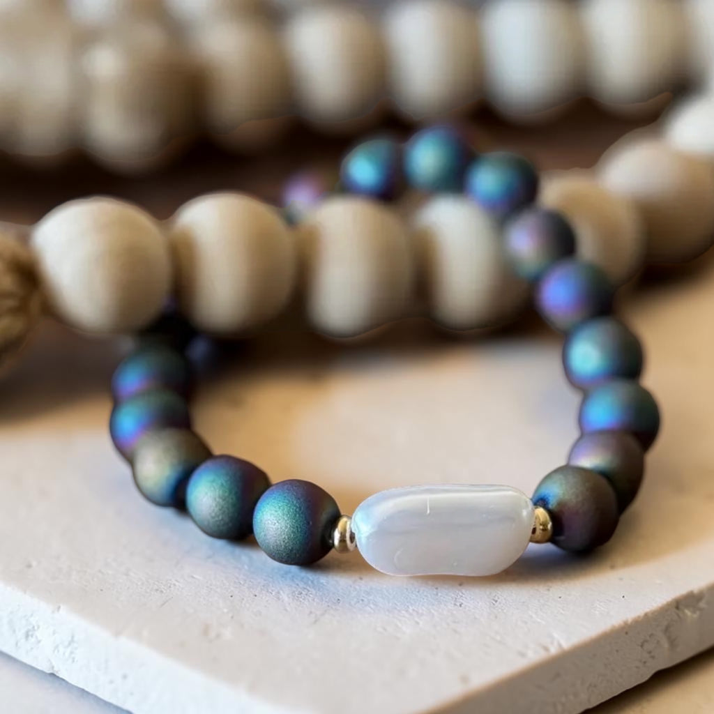 A beaded stretch bracelet featuring iridescent dark teal beads with a central white agate-like oval bead, displayed on a light stone surface with other blurred bracelets in the background.