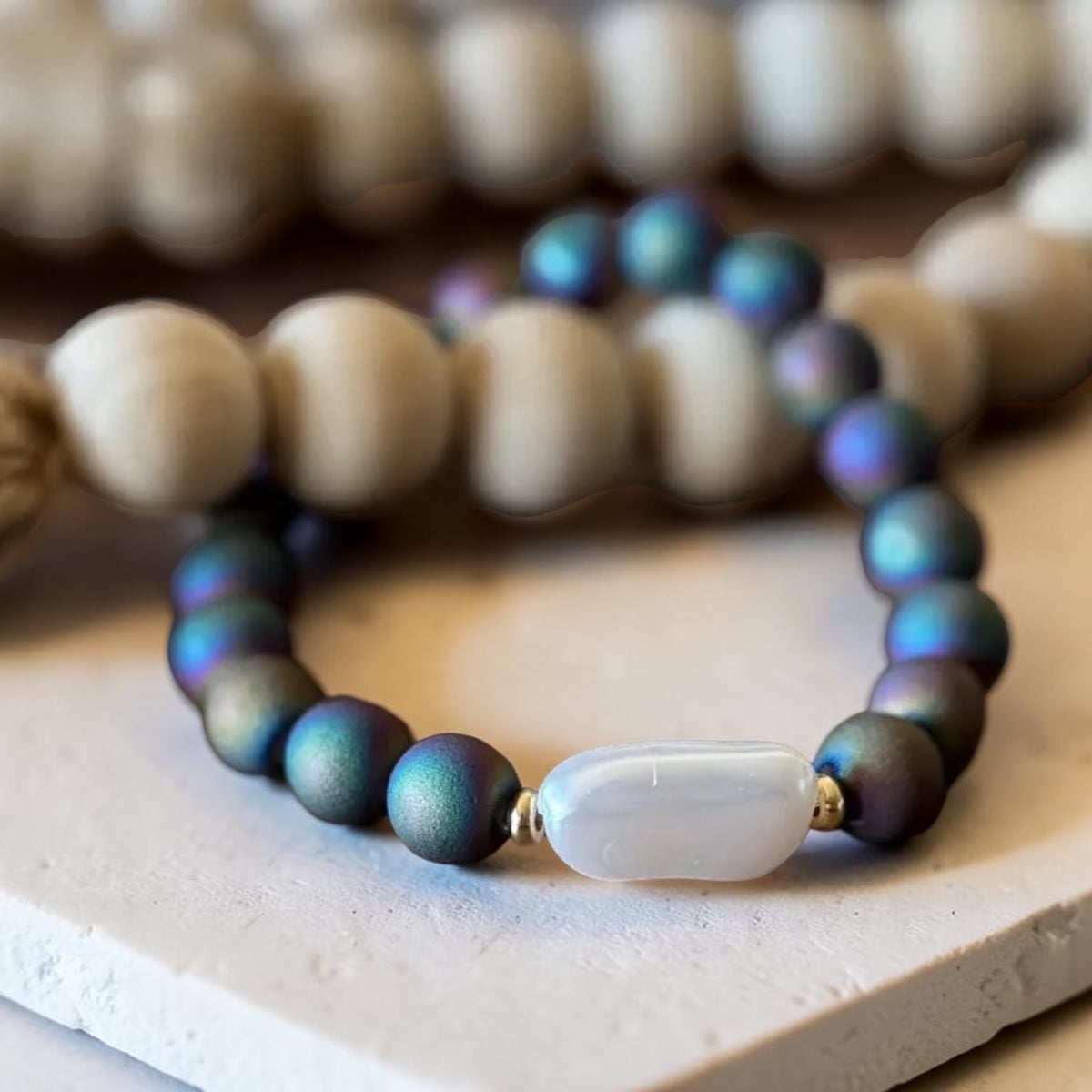 A beaded stretch bracelet featuring iridescent dark teal beads with a central white agate-like oval bead, displayed on a light stone surface with other blurred bracelets in the background.