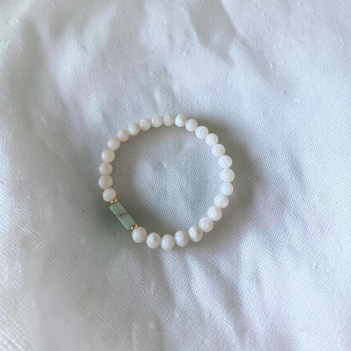 A beaded stretch bracelet with small white beads and a single light green stone accent, displayed on a white fabric background.