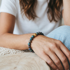 A close-up of a person's wrist wearing a beaded bracelet with iridescent blue-green beads and a small gold accent bead, while wearing a white t-shirt and light blue jeans.