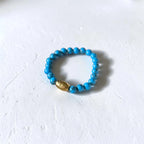 A beaded stretch bracelet featuring bright turquoise blue beads and a single textured gold accent bead, displayed on a textured white surface.