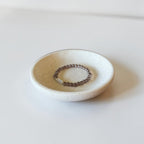 A small round ceramic dish holding a beaded stretch bracelet with dark brown beads and a small white stone accent, photographed on a clean white surface.
