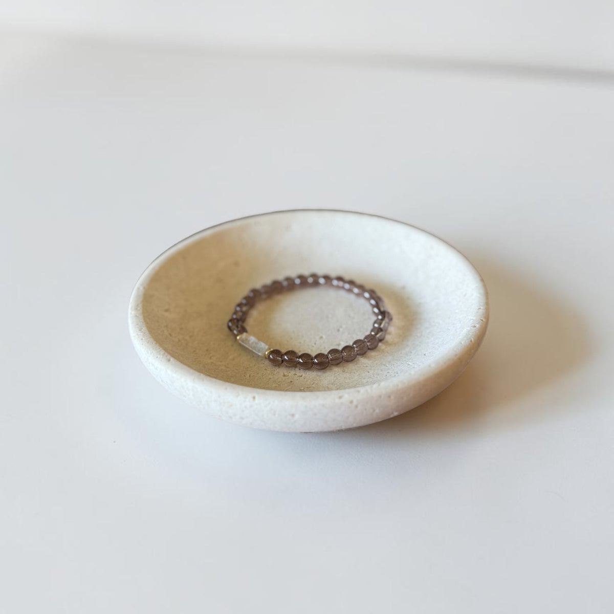A small round ceramic dish holding a beaded stretch bracelet with dark brown beads and a small white stone accent, photographed on a clean white surface.