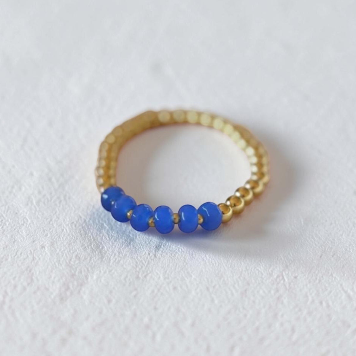 A gold beaded bracelet with five smooth blue beads on the front, photographed on a textured white surface.