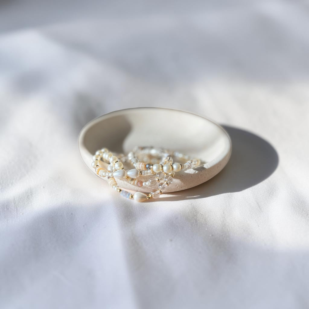 A small round ceramic dish holding a strand of white and cream beaded jewelry, photographed on a white fabric surface with soft natural shadows.