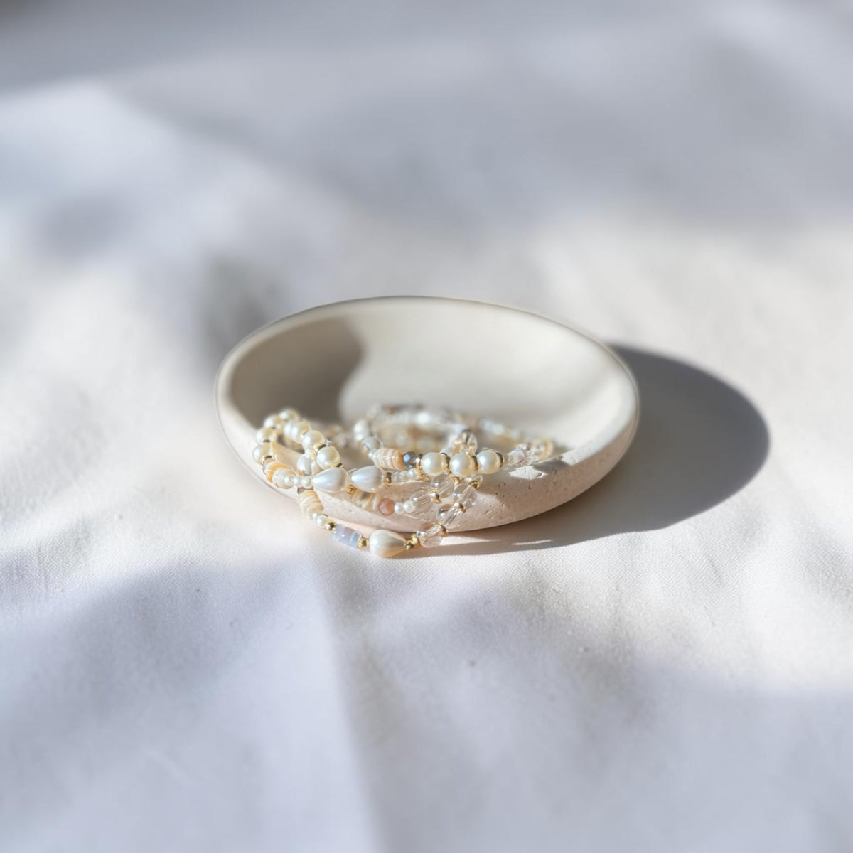 A small round ceramic dish holding a strand of white and cream beaded jewelry, photographed on a white fabric surface with soft natural shadows.