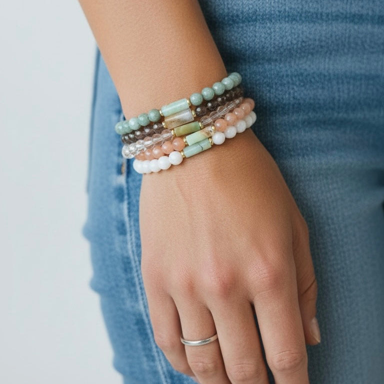 A close-up of a person's hand wearing a stack of five beaded stretch bracelets in pastel colors, paired with a silver band ring and blue jeans.