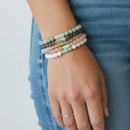 A close-up of a person's hand wearing a stack of five beaded stretch bracelets in pastel colors, paired with a silver band ring and blue jeans.