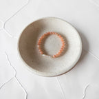 A peach-orange beaded bracelet with a silver-toned clasp resting inside a small, round, speckled beige ceramic dish on a white textured surface.