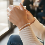 A close-up lifestyle shot of a person wearing a delicate white pearl bracelet on their wrist, holding a white ceramic coffee cup in a softly blurred indoor setting.