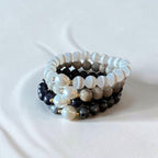 Three beaded stretch bracelets stacked together on a white textured fabric background. The bracelets feature white mother-of-pearl beads, gray agate beads, dark navy spiny oyster shell beads, and small gold spacer beads.