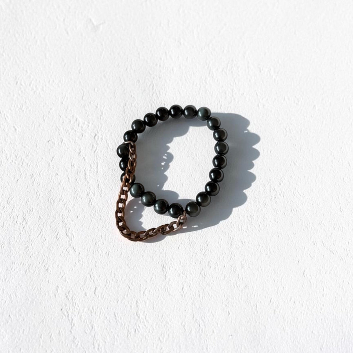 A beaded stretch bracelet featuring glossy black round beads and a short section of bronze-toned chain, photographed on a textured white background with a soft shadow.