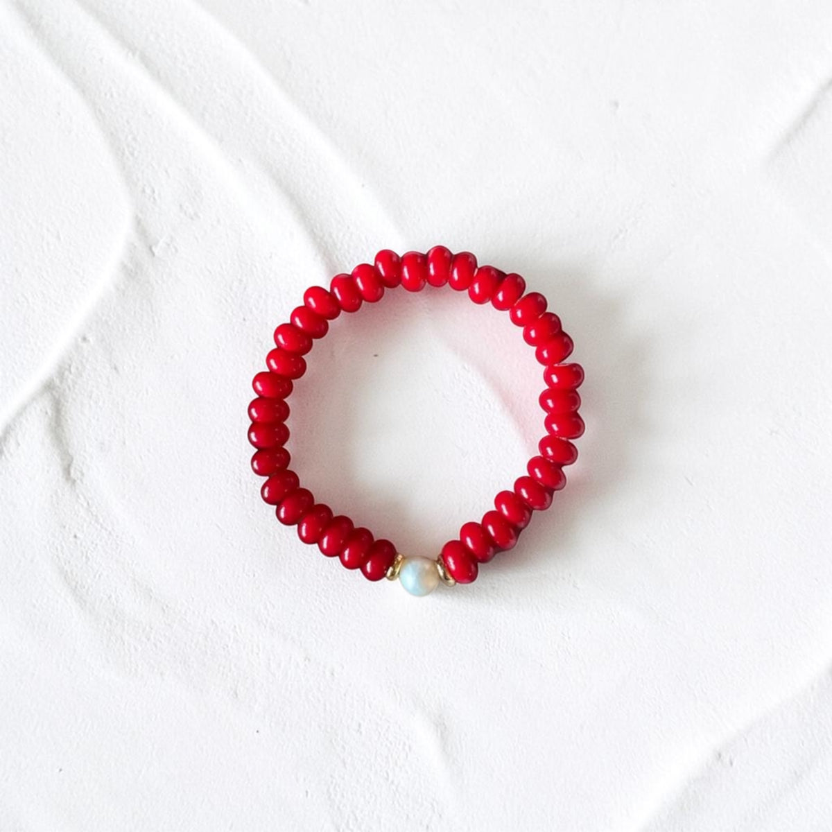 A red beaded stretch bracelet with a single white pearl accent bead, displayed on a textured white background.