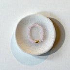 A pink beaded bracelet with a small gold accent bead, displayed inside a round, speckled ceramic dish on a textured off-white surface.