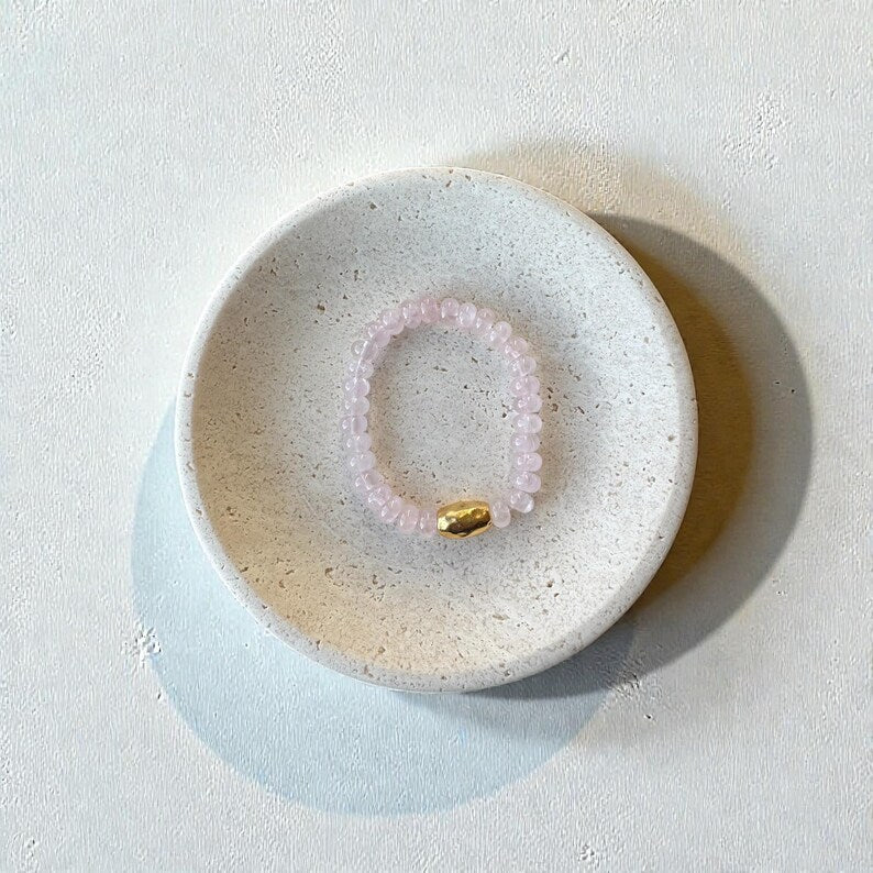 A pink beaded bracelet with a small gold accent bead, displayed inside a round, speckled ceramic dish on a textured off-white surface.