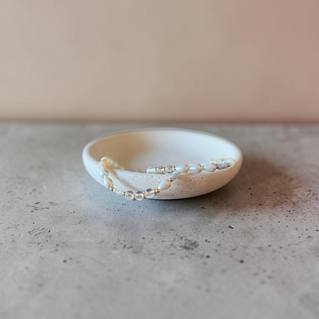 A delicate beaded bracelet with clear and white beads draped over a small, shallow off-white ceramic dish on a textured gray surface against a warm beige background.