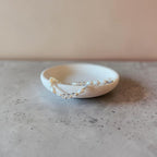 A delicate beaded bracelet with clear and white beads draped over a small, shallow off-white ceramic dish on a textured gray surface against a warm beige background.