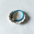 A beaded bracelet in shades of blue and teal with a small white pearl accent, displayed on a small white ceramic dish against a light gray textured background.