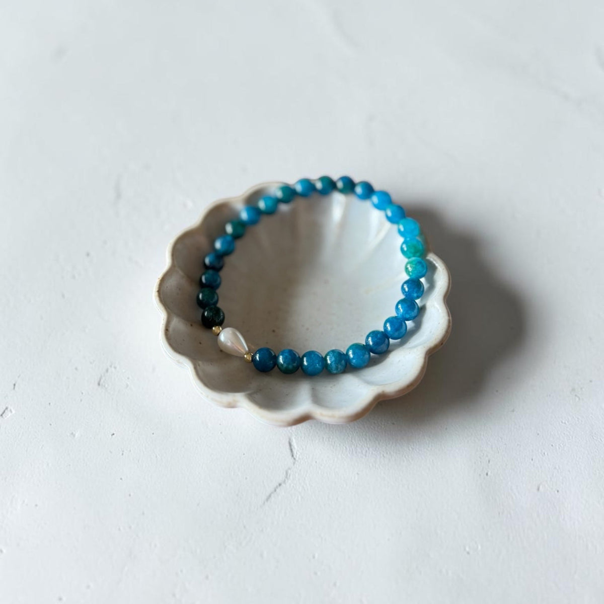 A beaded bracelet in shades of blue and teal with a small white pearl accent, displayed on a small white ceramic dish against a light gray textured background.