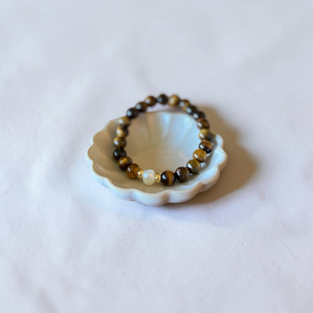 A beaded stretch bracelet with brown and black tiger's eye beads and a single white pearl, displayed on a small white ceramic shell-shaped dish against a soft white fabric background.