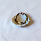 A beaded stretch bracelet with brown and black tiger's eye beads and a single white pearl, displayed on a small white ceramic shell-shaped dish against a soft white fabric background.