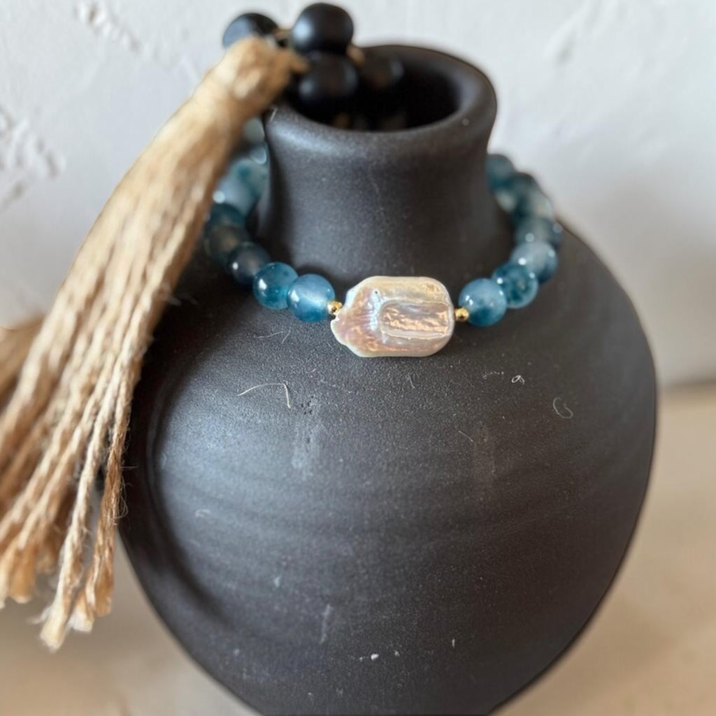 A beaded bracelet with blue faceted beads, a textured beige stone centerpiece, and a natural jute tassel, displayed on a matte black ceramic vase.