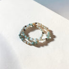 A pair of beaded stretch bracelets with light blue, white, and brown beads, arranged in a double loop on a textured white surface with soft shadows.
