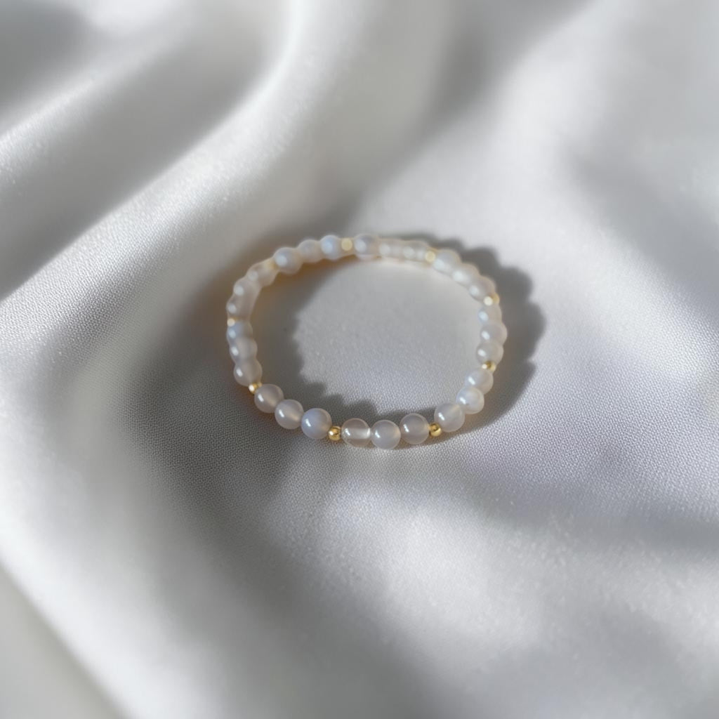 A beaded stretch bracelet made of small white pearl-like beads separated by tiny gold spacer beads, resting on draped white satin fabric.