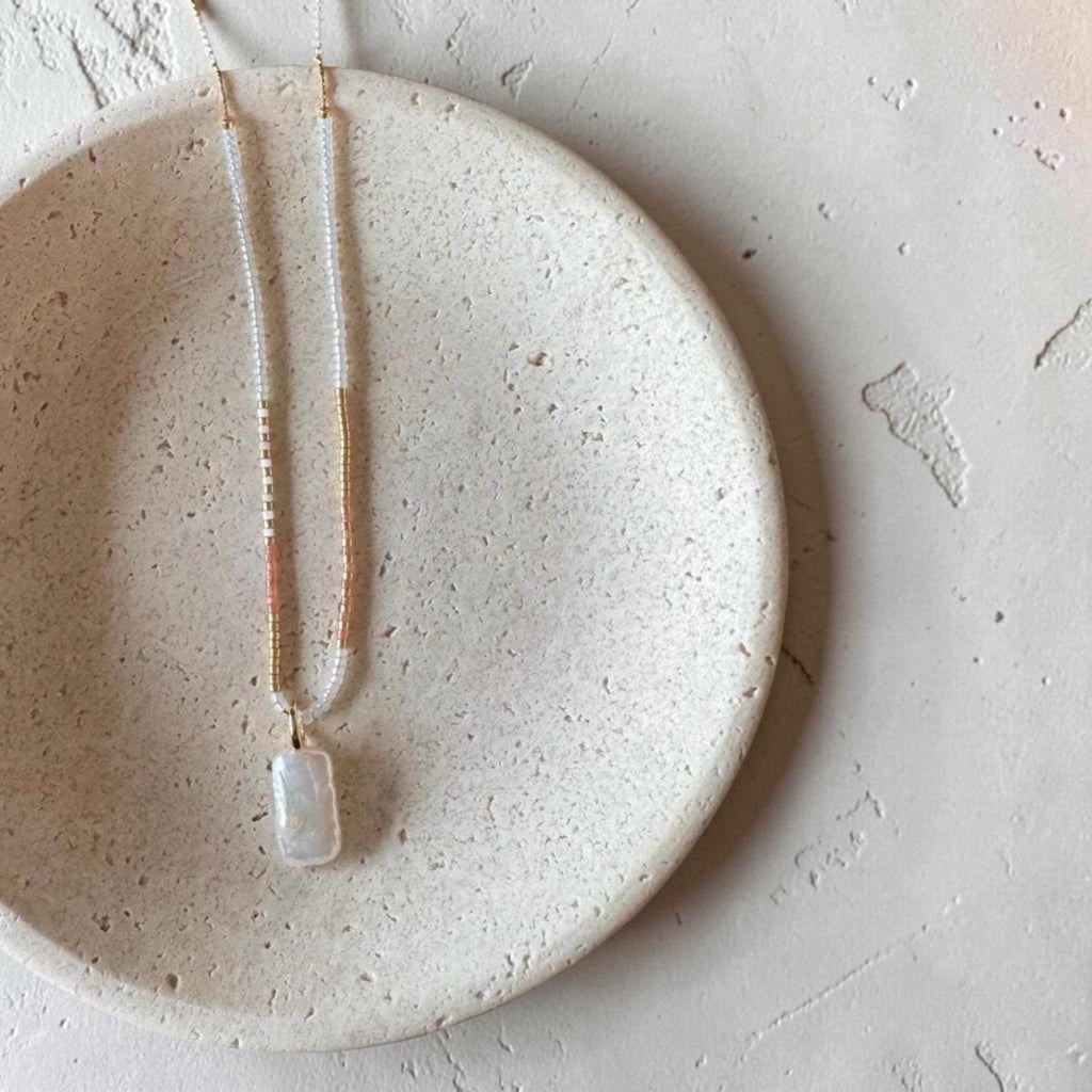A delicate necklace with a white rectangular pendant and gold-toned chain, displayed on a round beige ceramic dish against a textured light background.
