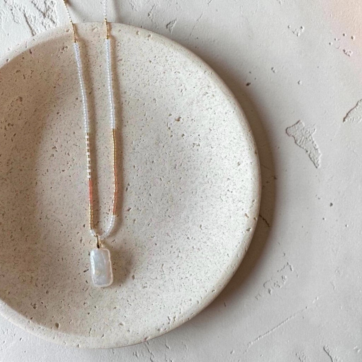 A delicate necklace with a white rectangular pendant and gold-toned chain, displayed on a round beige ceramic dish against a textured light background.